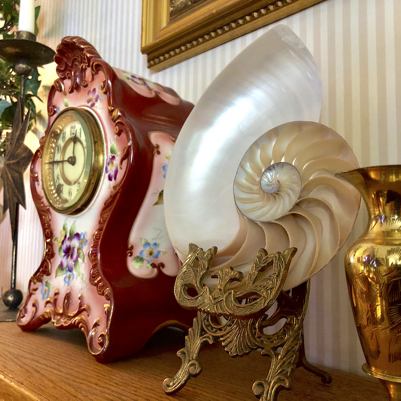 An antique clock, Victorian era decorative gilt shell, and a gold-tone vase decorate a shelf inside an antique store.